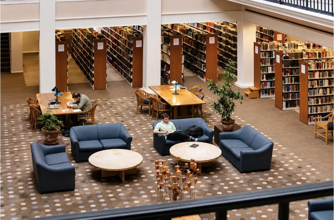 Library reading room with blue sofas and round tables on a dotted patterned carpet, seen from a balcony