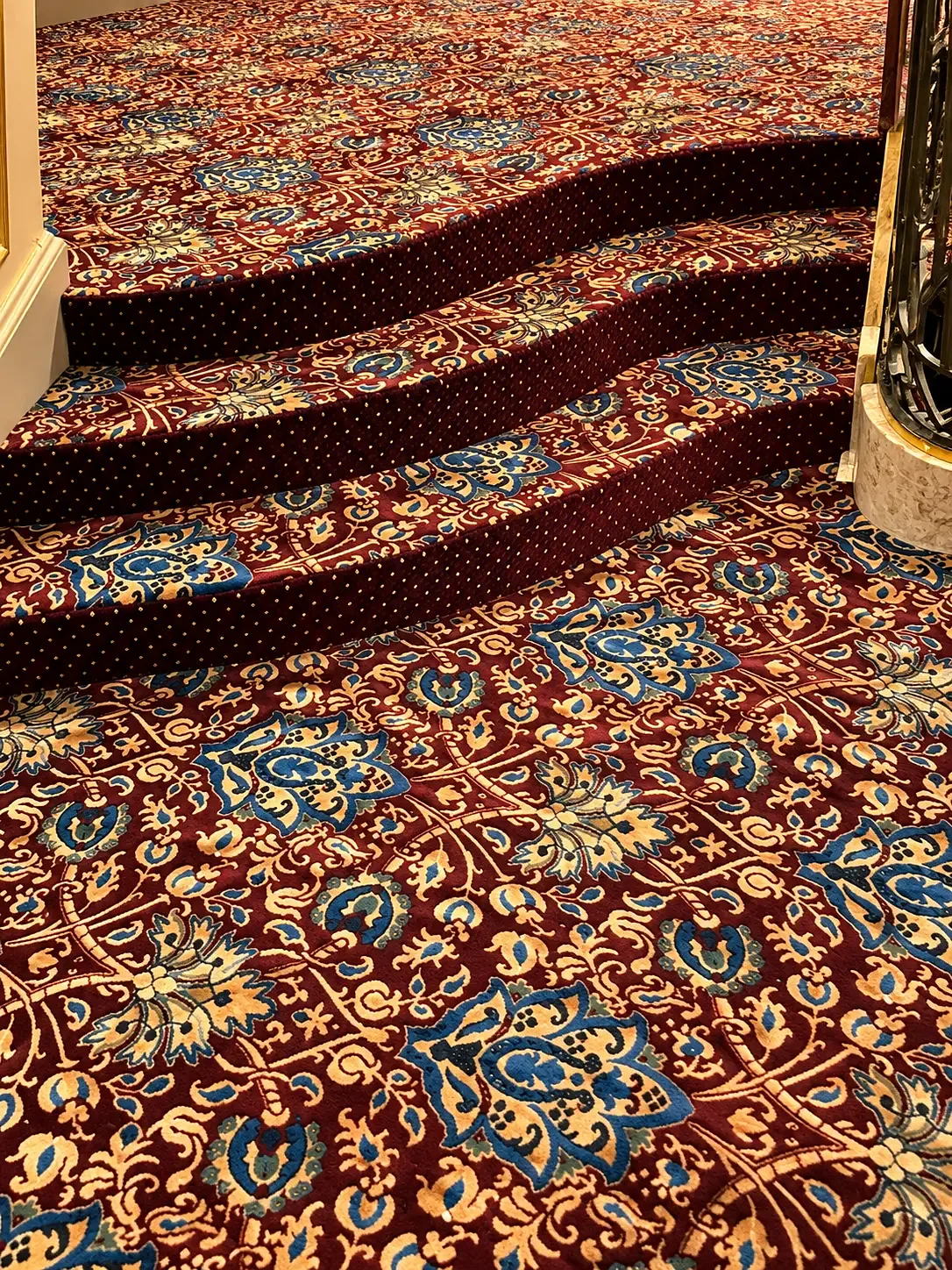 Close view of curved carpeted steps in an ornate red, blue and gold floral pattern with dotted risers